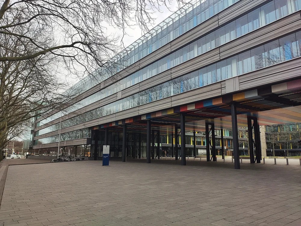 Een modern kantoorgebouw met een glazen gevel en een luifel met kleurige verticale stroken, gezien vanaf de straat met kale bomen op de voorgrond.