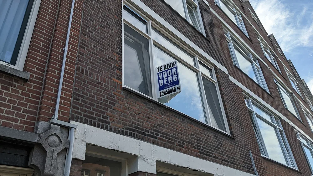 Een blauw-wit bord met de tekst 'TE KOOP' bevestigd aan het kozijn van een raam op de eerste verdieping van een bakstenen woonhuis, van onderaf gezien tegen een blauwe lucht.