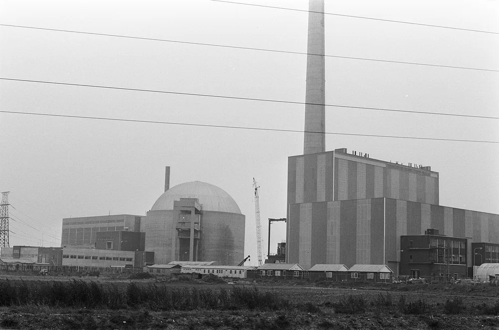 Zwart-witfoto van een industriecomplex achter een kaal veld: links een ronde koepel met een aanbouw, rechts een groot rechthoekig gebouw met verticale strepen en een hoge schoorsteen, op de voorgrond hoogspanningsdraden.