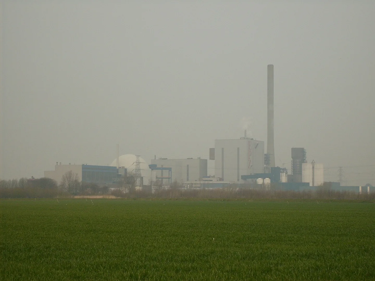 In de nevel achter een groen grasveld staan lichte fabrieksgebouwen, een witte koepel en een hoge schoorsteen; op een van de gebouwen staan rode letters EPZ.