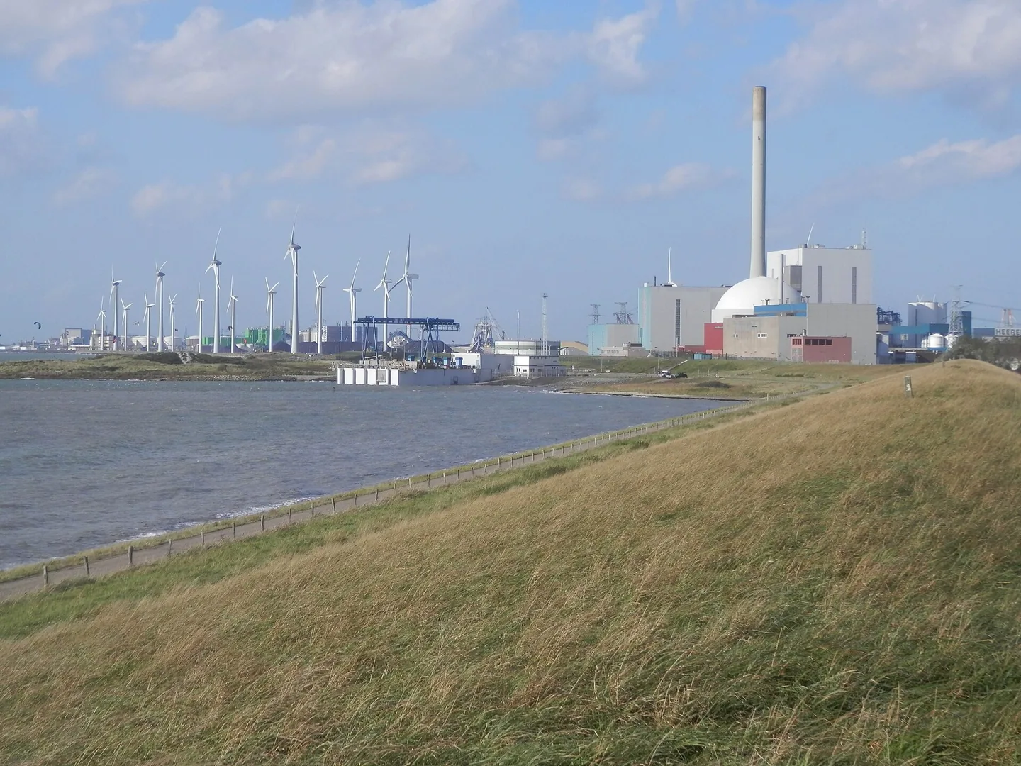 Een grasdijk loopt naar rechts omhoog; daarachter water en een industrieterrein met een witte koepel, een hoge schoorsteen en lichte fabrieksgebouwen, links een rij windturbines onder een blauwe lucht met wolken.