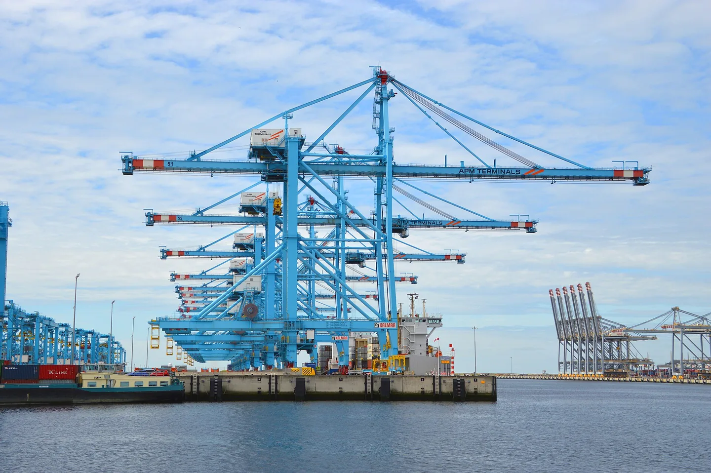 Een rij lichtblauwe containerkranen aan een kade, gezien vanaf het water, met verderop nog meer kranen onder een lichtbewolkte lucht.