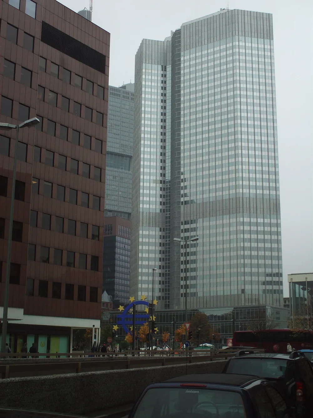 Een grijze kantoortoren achter een drukke straat met auto's; onderaan staat een blauw euroteken omringd door gele sterren.