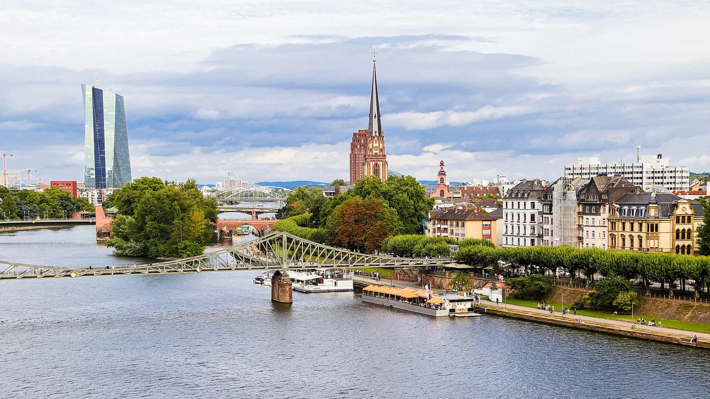 Een brede rivier met een ijzeren vakwerkbrug en rondvaartboten; langs de rechteroever staan oude gevels, in de verte links een glazen wolkenkrabber en midden een rode kerktoren.
