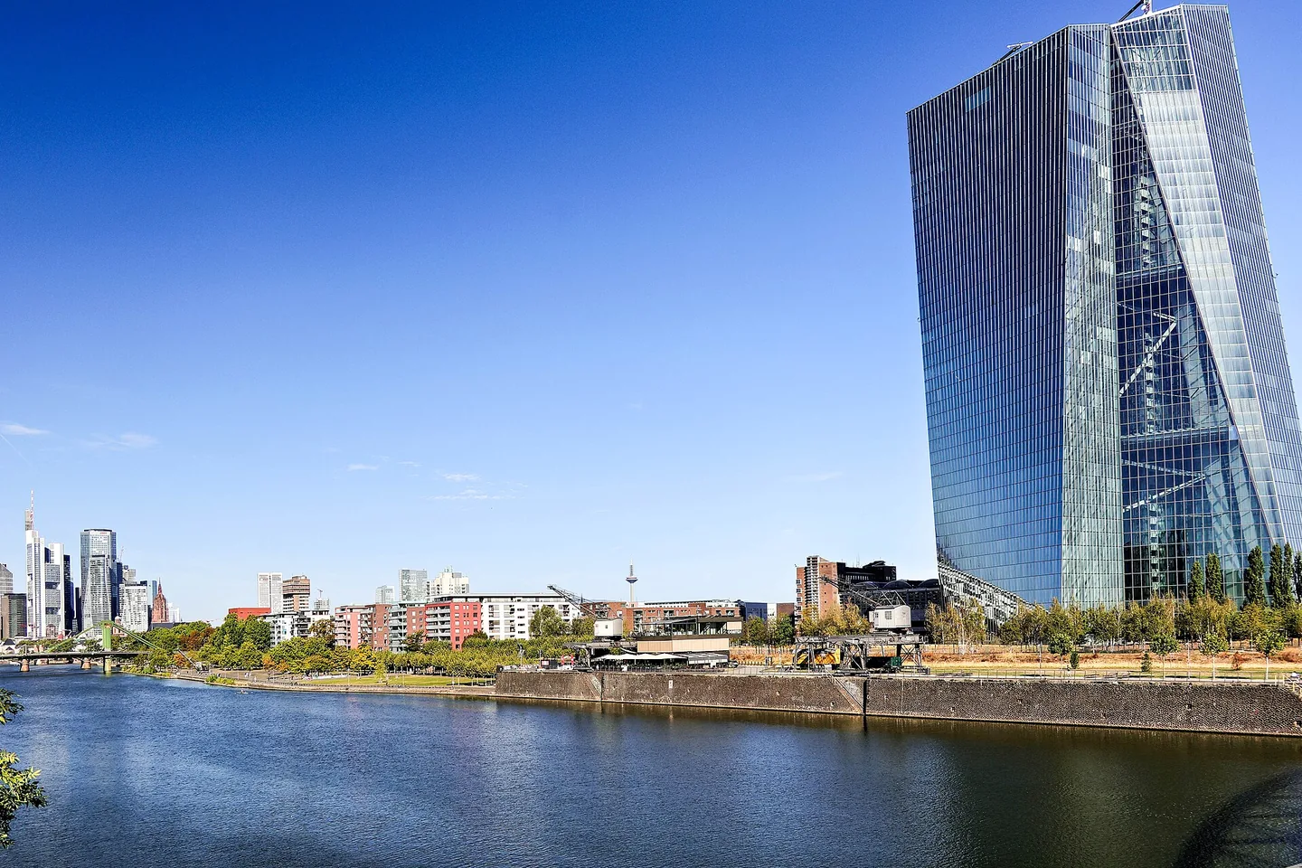 Een hoge glazen toren met twee schuin tegen elkaar geplaatste delen staat rechts aan een rivierkade onder een strakblauwe lucht; links aan de horizon staan wolkenkrabbers.