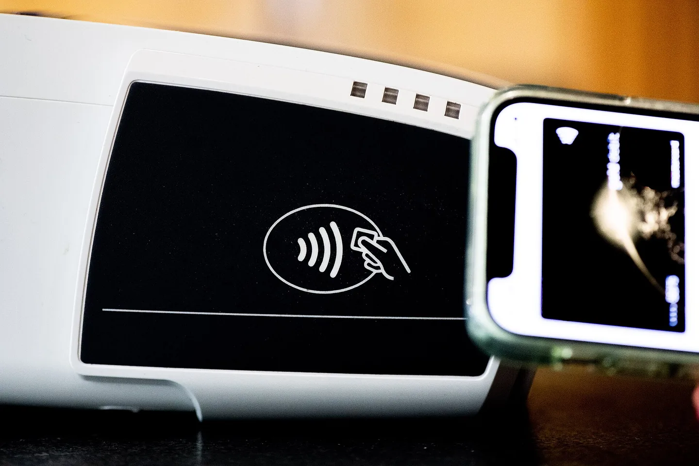 Close-up van een witte betaalterminal met een zwart vlak en het symbool voor contactloos betalen; rechts een smartphone met een betaalkaart op het scherm.