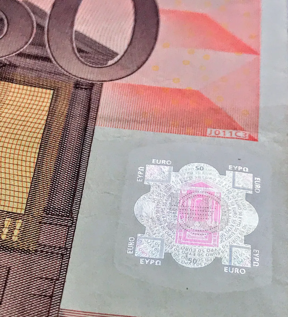 Close-up van de hoek van een oranjebruin biljet van 50 euro met een glanzend zilveren vlak waarin een poortje en de woorden EURO en ΕΥΡΩ te zien zijn.