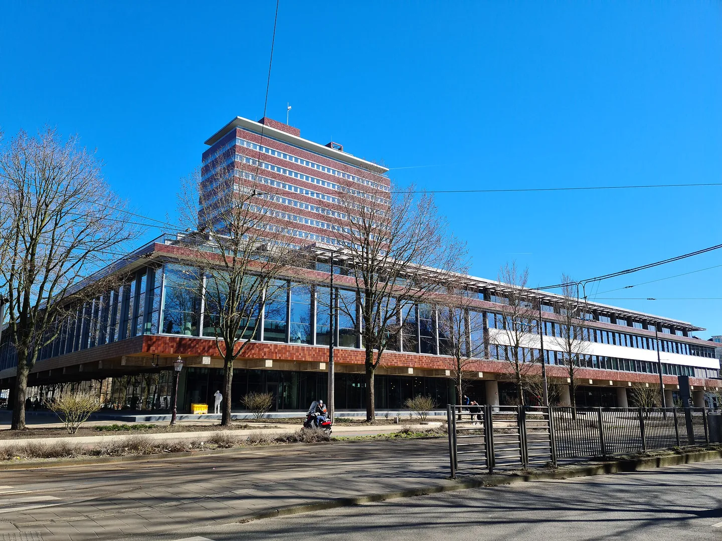 Een breed modern kantoorgebouw met glazen gevels en roodbruine banden, met erachter een hoge toren, onder een strakblauwe lucht met kale bomen op de voorgrond.