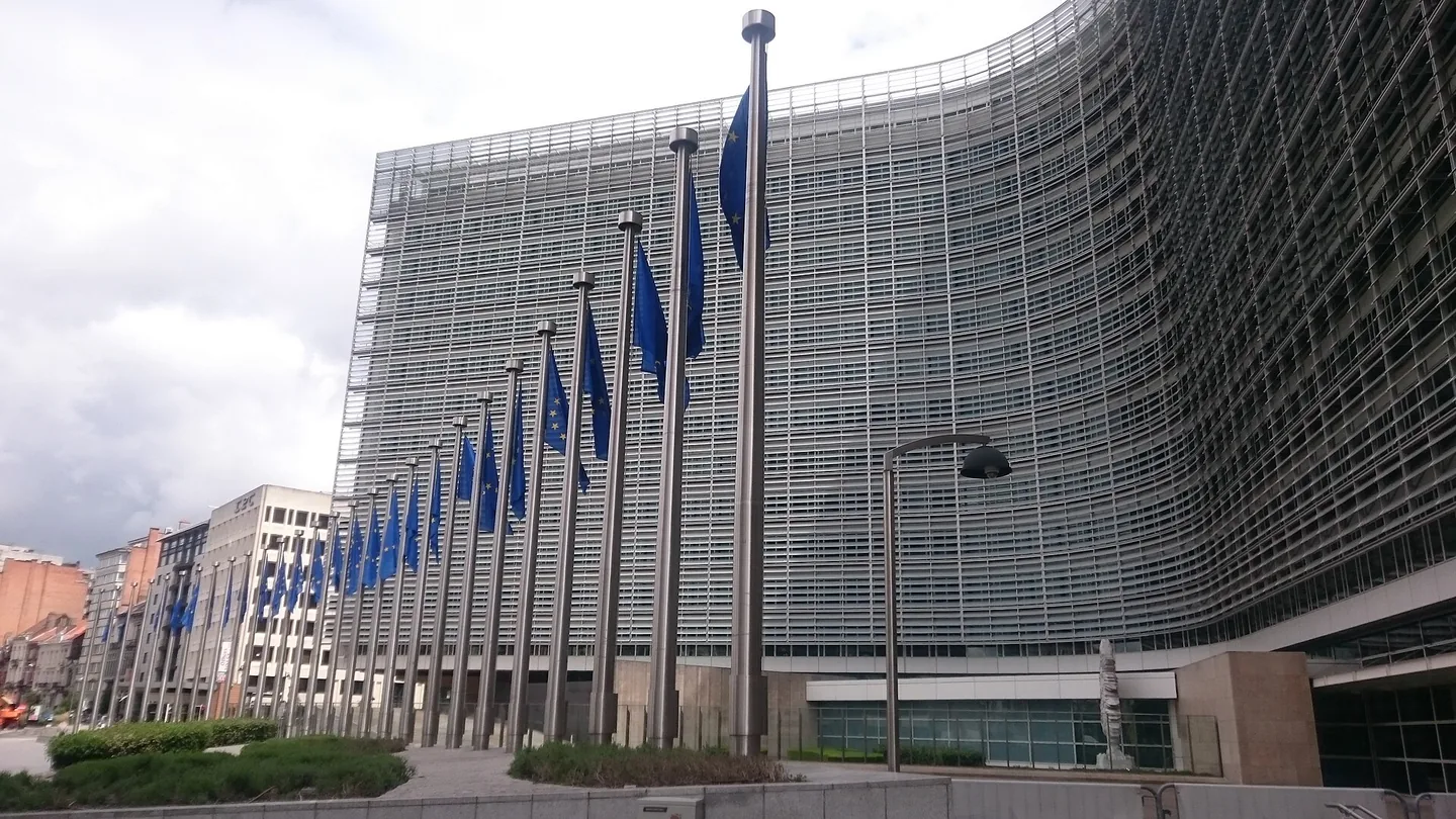 Een gebogen glazen kantoorgebouw met een lange rij vlaggenmasten waaraan blauwe Europese vlaggen wapperen, onder een bewolkte lucht.
