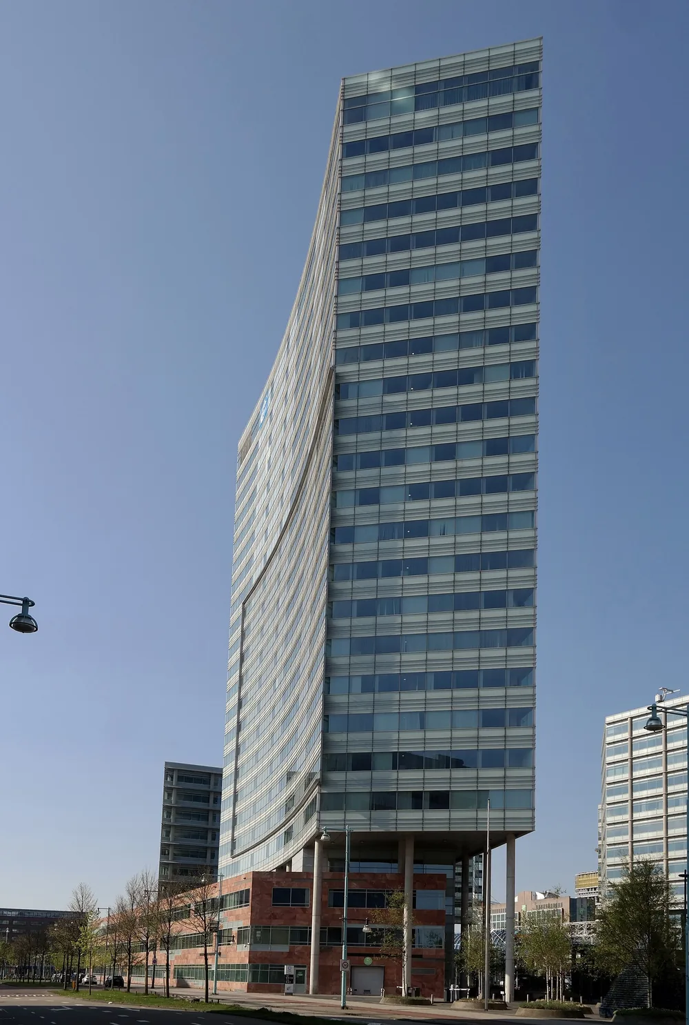 Een smalle glazen kantoortoren op palen met een gebogen zijgevel, tegen een strakblauwe lucht.