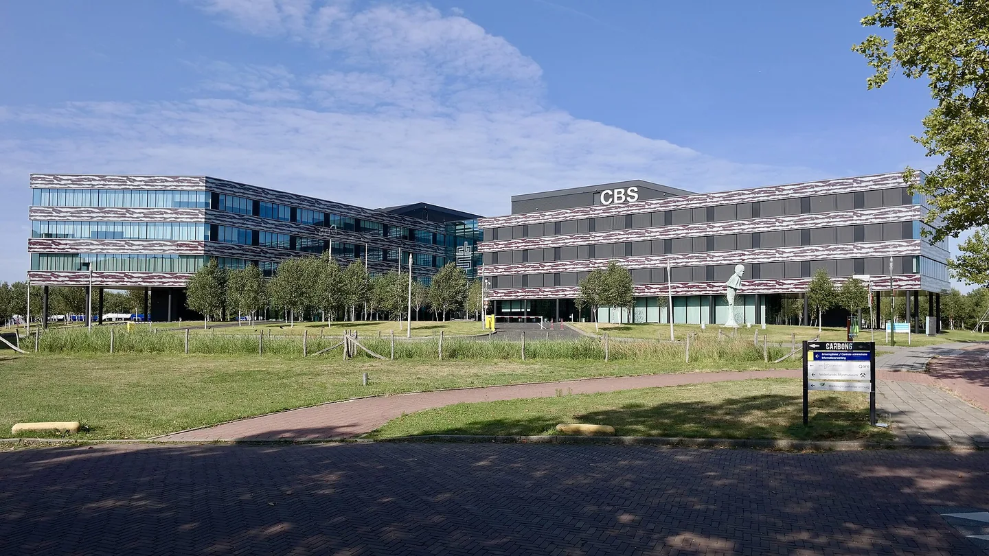 Twee lage kantoorgebouwen met gestreepte gevels en de letters CBS op het dak, achter een grasveld met een klinkerpad.