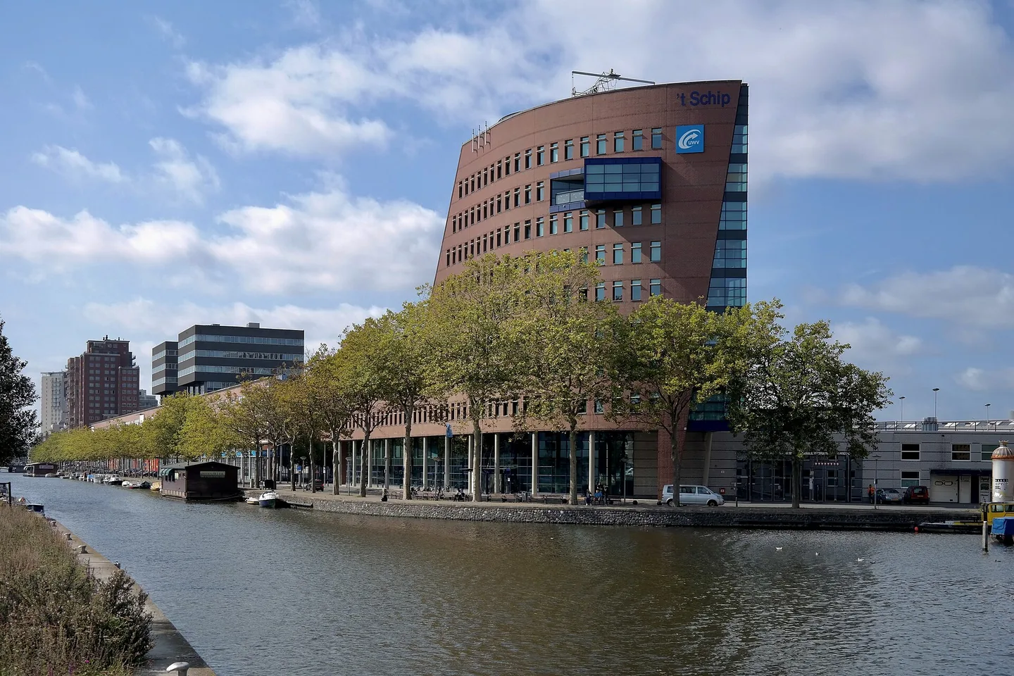 Een roodbruin kantoorgebouw met een gebogen voorkant aan een gracht met bomen langs de kade; op de gevel staan de naam 't Schip en het logo van UWV.
