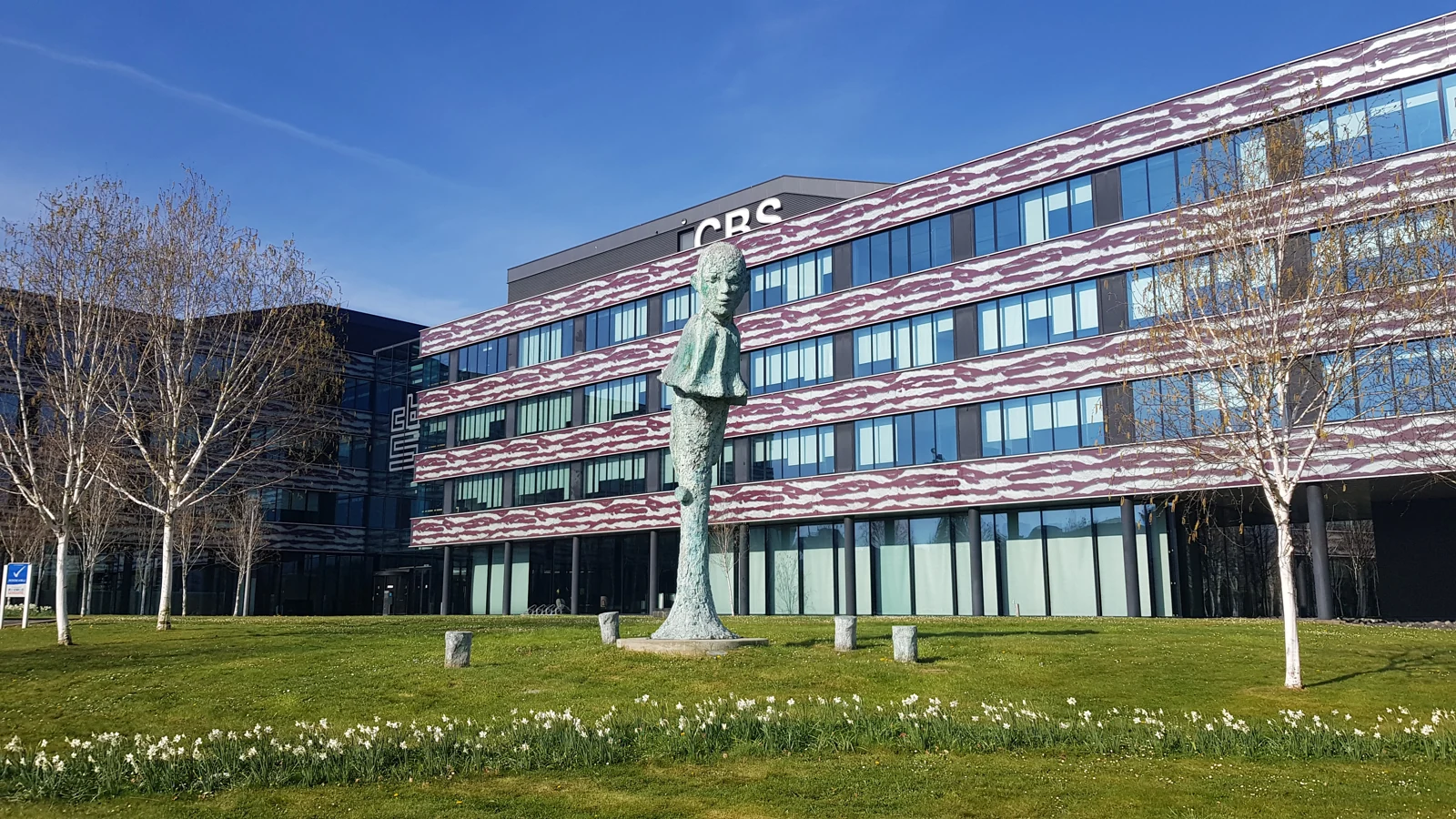 Het glazen CBS-kantoorgebouw in Heerlen, met bomen en een grasstrook.