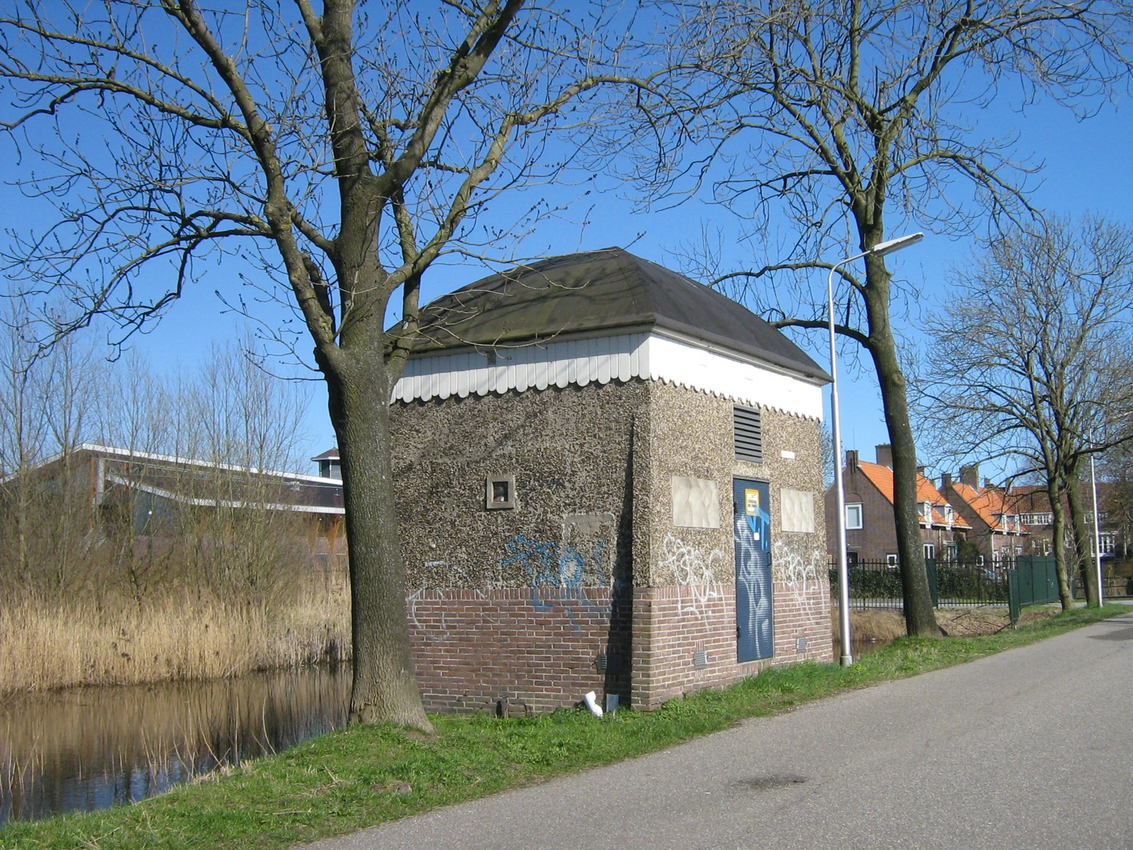 Klein bakstenen elektriciteitsgebouw tussen bomen langs een weg.